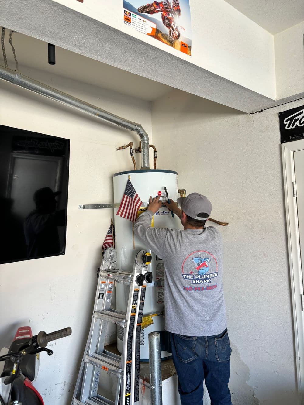 Plumber installing water heater with American flags in garage setting. Tools and ladder visible.
