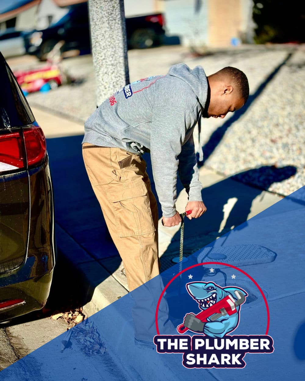 Plumber unclogging a drain outside, wearing a grey hoodie and tan pants, The Plumber Shark logo.