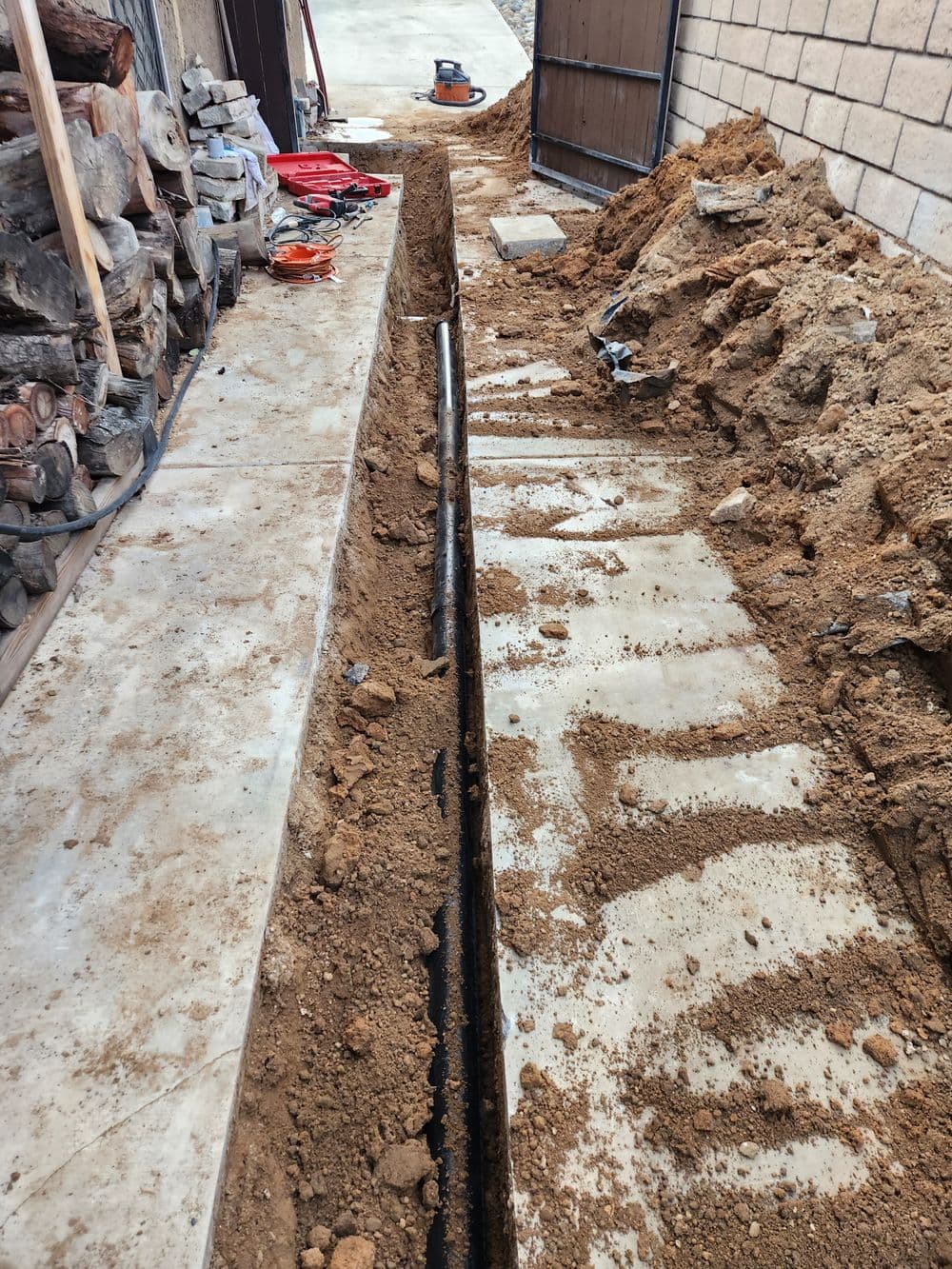 Excavated trench for utility installation beside a building with wood piles nearby.