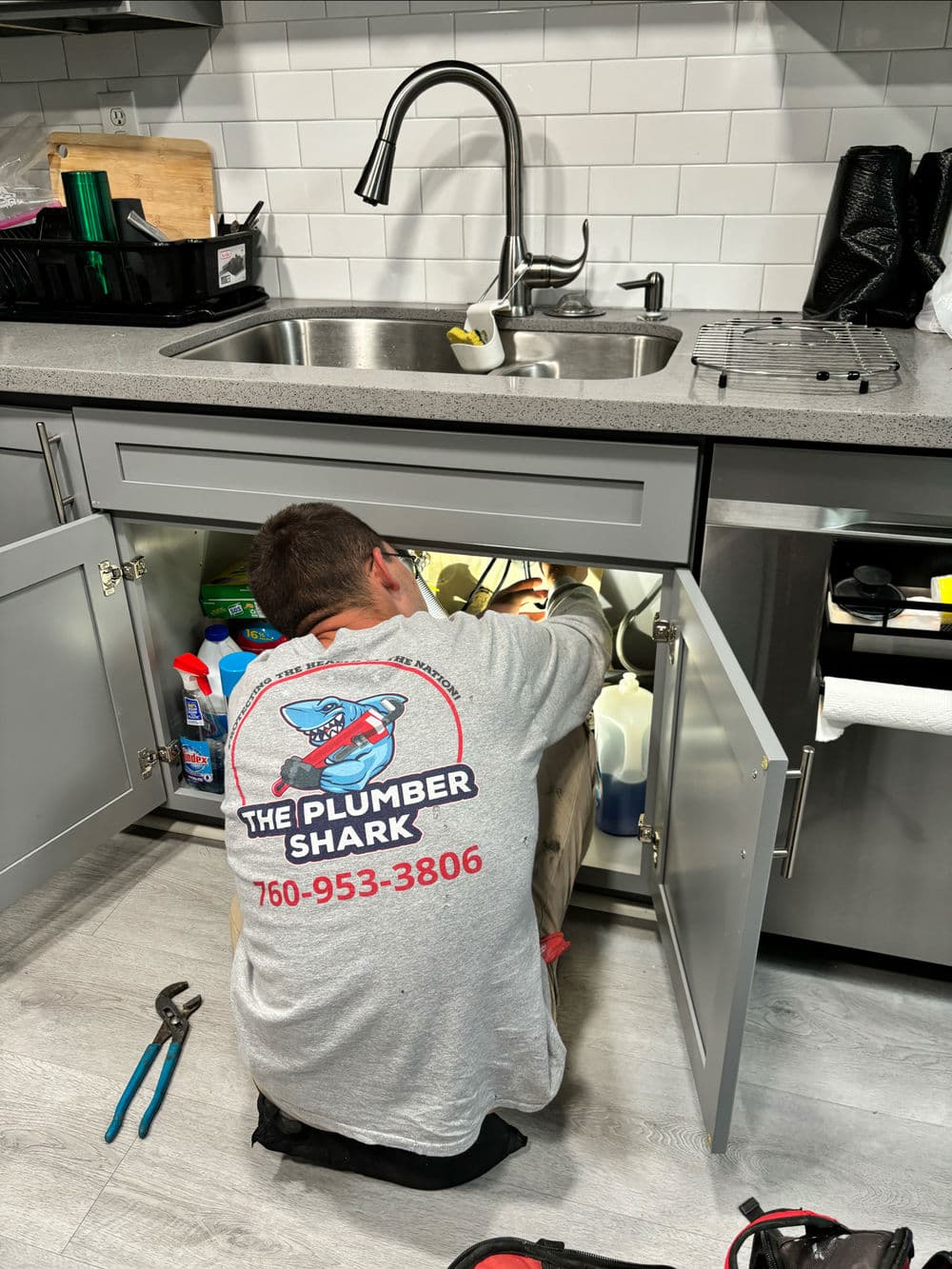 Plumber in gray shirt fixing sink under kitchen cabinet with cleaning supplies.
