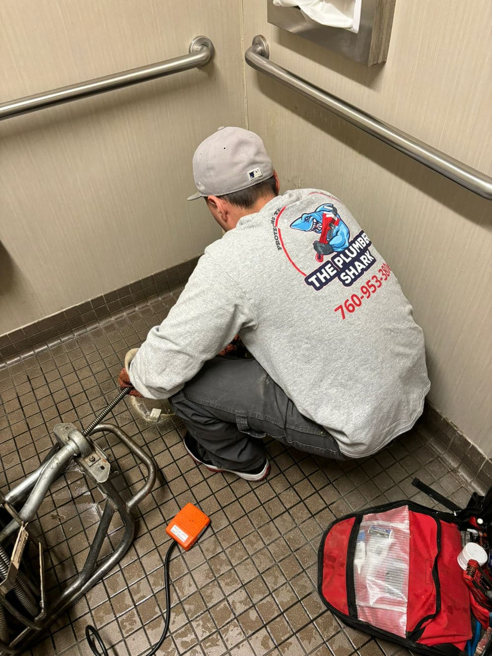 Plumber repairing drainage in commercial restroom, tools and equipment visible on floor.